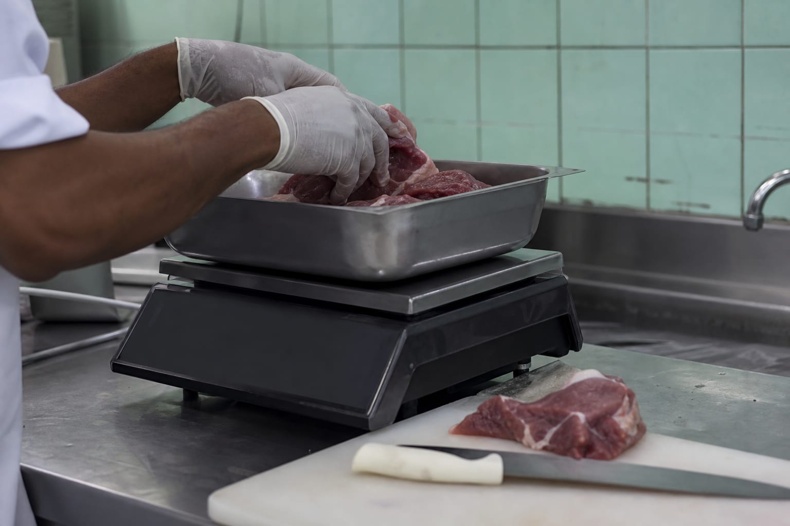 Mãos com luva pesam carne em cuba de aço sobre balança de bancada de cozinha industrial